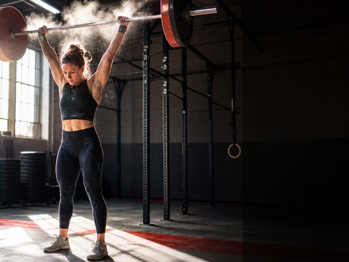 An athlete at lockout under a loaded barbell overhead, chalk bursting off the bar into the light.
